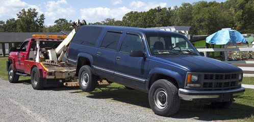 Red tow truck hauling blue vehicle in rural setting. Horizontal.