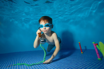 Little boy underwater swims and plays in the pool with the handset. Portrait. Shooting underwater. Landscape orientation - obrazy, fototapety, plakaty
