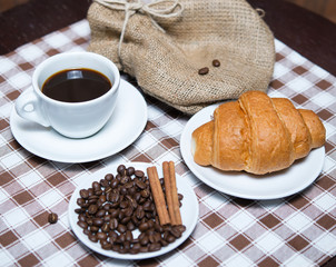 fresh black coffee with beans and croissant on tablecloth