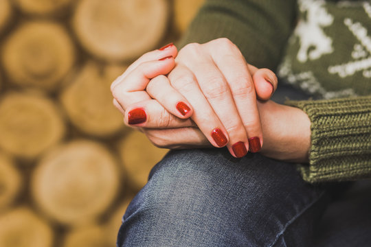 Woman sitting indoors with her hands crossed on knee. Young woman wears green sweater and blue denim jeans. Fingernails with beautiful fresh red manicure. Close up horizontal color photo.