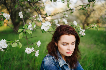 Naklejka premium Young pretty serious caucasian woman in jeans jacket standing near blooming spring tree. Youth, freshness, beauty, happiness concept.