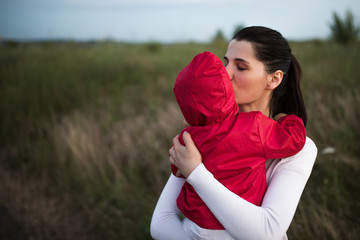 Beautiful young mother holding and kissing her baby toddler wearing red jacket. Happy family, mother and child play cuddling on field walk in nature outdoors.