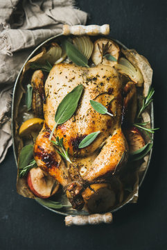 Oven roasted whole chicken with onion, apples and sage in serving tray over dark stone background, top view, selective focus, vertical composition. Celebration food concept
