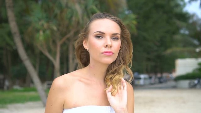 Pretty blond woman in sexy white summer dress walking at the beach and smiling at the camera against beautiful green trees tropical background - video in slow motion