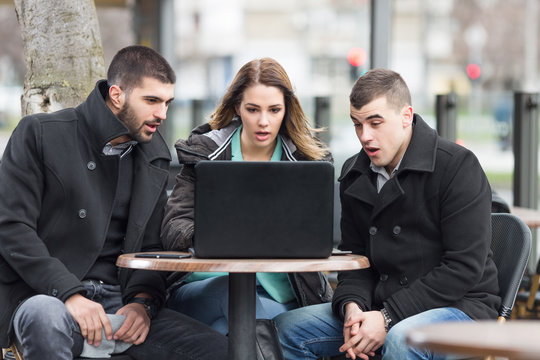 Group of students or young business people is sitting in an outdoor caffe and using laptop. They are suprised.