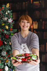 Portrait of beautiful redhead woman holding plate with christmas cakes before dressed up tree