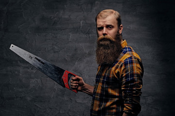 A bearded carpenter man dressed in a plaid shirt holds handsaw.