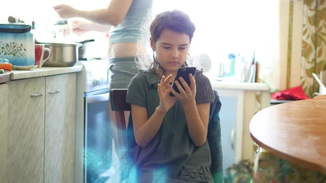Girl in the kitchen playing smartphone rear mother preparing breakfast