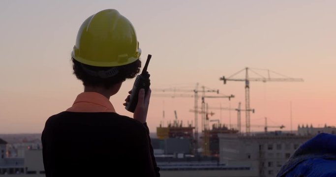 Woman wearing hard hat and smart clothes of architect, building supervisor or contractor, talks on walkie talkie with cranes in background in Downtown Los Angeles. Medium close up from rear at sunset