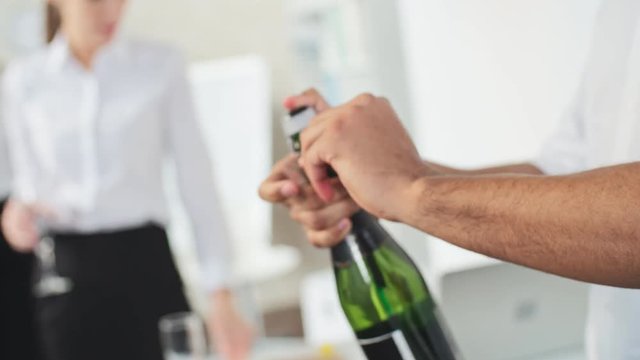 Man opening champagne bottle and pouring drink into flutes of colleagues celebrating holiday in the office