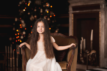 Smiling baby girl 4-5 year old wearing trendy white dress sitting in vintage chair in room over Christmas tree with lights. Looking at camera. Holiday season.