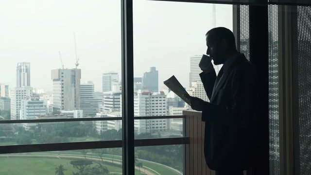 Young businessman reading magazine and drinking coffee by window in office
