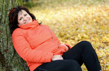 Obraz premium Portrait of smiling middle aged ordinary caucasian woman with long dark hair and red jacket leaning on tree.