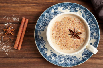 Masala tea chai latte traditional hot Indian sweet milk with spices, cinnamon stick, herbs blend organic infusion healthy beverage in porcelain cup on wooden table background