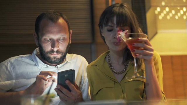 Bored couple sitting in cafe, man using smartphone, woman drinking cocktail
