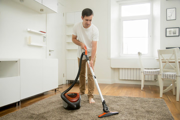 Happy smiling man vacuum cleaning the carpet in the living room, modern scandinavian interior....