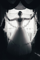 silhouette of a bride is standing at the window and holding a veil. black and white portrait of a...