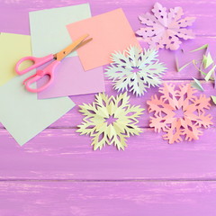 Colorful paper snowflakes ornaments, colored paper sheets and scrap, scissors on lilac wooden background. Making snowflakes out of colored paper. Easy winter crafts project for kids