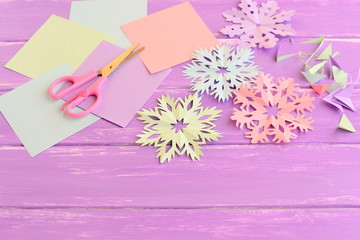 Colorful paper snowflakes, colored paper sheets and scrap, scissors on lilac wooden background. Cutting snowflakes out of colored paper. Simple winter crafts idea for children