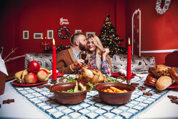 Beautiful couple kissing and holding glass of wine in a decorated festive interior with a Christmas tree. A romantic dinner for thanksgiving with fried chicken and candles