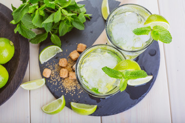 Two fresh mojitos cocktail on wooden background. Mojitos with mint leaves, lime and ice. Drink making tools and ingredients for cocktail. View from above.  