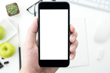 Hand is holding a smartphone with blank mockup screen above the white desk table.