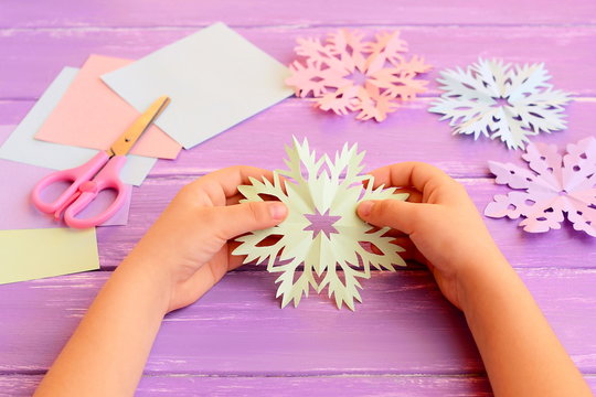 Child holds a paper snowflake in hands. Child shows snowflake decoration. Colored paper, scissors, snowflakes crafts on wooden background. How to make simple structures from a paper. Kids winter art