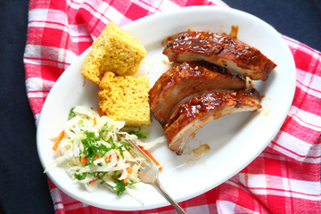 Overhead view of barbecued ribs with coleslaw and cornbread with a red-and-white checked cloth
