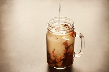 Pouring milk into a jug with delicious iced coffee on grey background