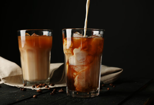 Pouring cream into a glass with iced coffee on wooden table and black background