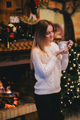 Beautiful young woman sitting near fireplace under the Christmas tree drinking cocoa with marshmallow. Cozy mood