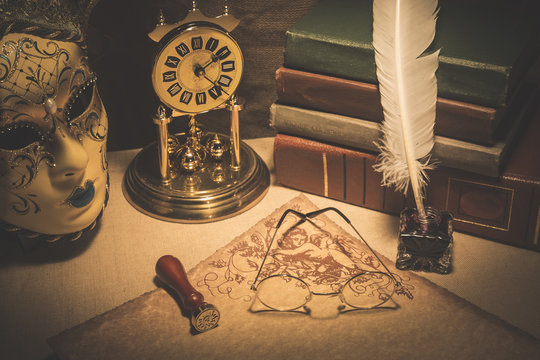Old glasses on scroll with books, old clock, feather in inkstand and venezian mask. Vintage toned photo