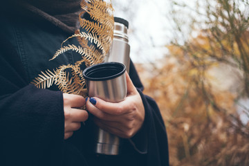 Woman traveler hands holding thermos bottle and cup of tea, walking in autumn forest. Adventure, travel, tourism and camping concept. - obrazy, fototapety, plakaty