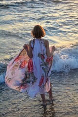 Mädchen von hinten steht im Wasser am Strand, hält mit Händen langes Kleid nach oben 