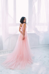 Beautiful brunette girl standing in a bright room. She is dressed in luxurious, fluffy, pink dress. Gentle puppet's face. The European image of the bride. Photo high key style.