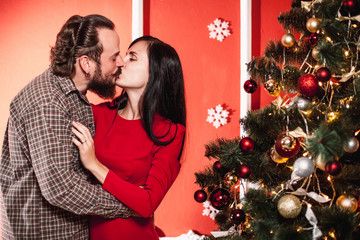 Family portrait in home holiday living room, loving young couple hugging and kissing near the Christmas tree in decorated New Year interior
