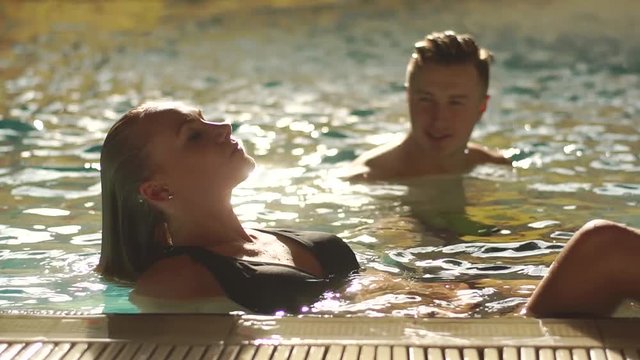 Attractive couple swimming in the pool in european hotel. Man is looking exciting at girl. Blonde woman on the edge of the pool taking her head back down and wetting the hair .