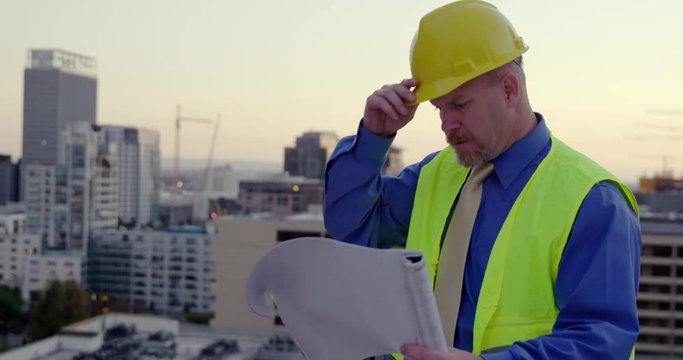 Man wearing hard hat and safety vest over smart clothes of architect, building supervisor or contractor, looks at plans and scratches his head in Downtown Los Angeles. Hand held, MS recorded at 60fps