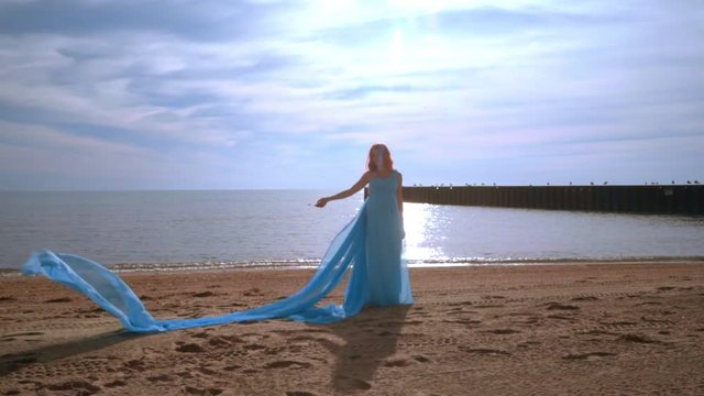 Pregnancy fashion. Pregnant model in blue dress on beach. Woman pregnant on beach sea. Man throwing fabric on wind. Pregnant woman in blue dress flying on wind at beach. Romantic photo session