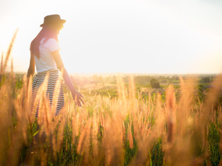 Trendy girl in stylish summer dress with beautiful hat walking in the field with flowers in sunlight