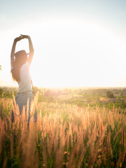 Trendy girl in stylish summer dress with beautiful hat walking in the field with flowers in sunlight