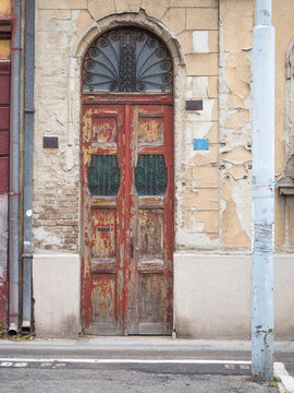 Picture of the front door with wrought iron grill. Some of the red paint has scaled off. An old building with cracky beige - coloured paint and rusty gutter. Background of the old building