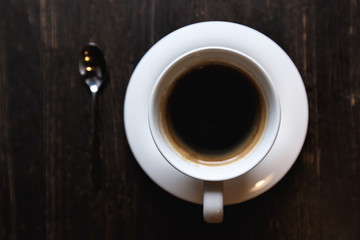 A dark, soft-focus image of a white coffee cup with black coffee and a tea spoon on a dark wooden table