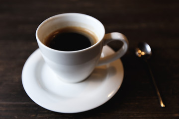 A dark, soft-focus image of a white coffee cup with black coffee and a tea spoon on a dark wooden table