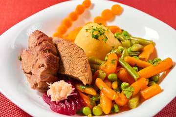 Traditional Romanian calf boiled meat with boiled vegetables on white plate and red place mat, tilted view