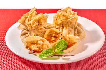 Traditional Romanian crepes with banana slices, hazelnut spread with cocoa and mint leaves decoration, on white plate and red place mat, isolated on white background