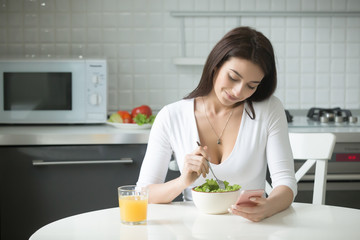 Portrait of happy attractive woman sitting at the kitchen table, having healthy lunch, salad and orange juice, holding smartphone