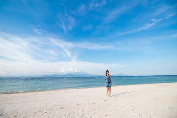 Fototapeta na wymiar Young woman in blue dress walking on beach of tropical island Nusa Lembongan, Indonesia. Amazing sky, ocean view. Volcano Agung and island Bali on the background.