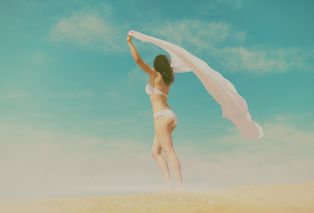 Young woman in bikini with white pareo walking against blue sky with clouds back to camera