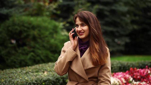 Young beautiful brunette woman in beige coat sitting on a bench in the autumn park